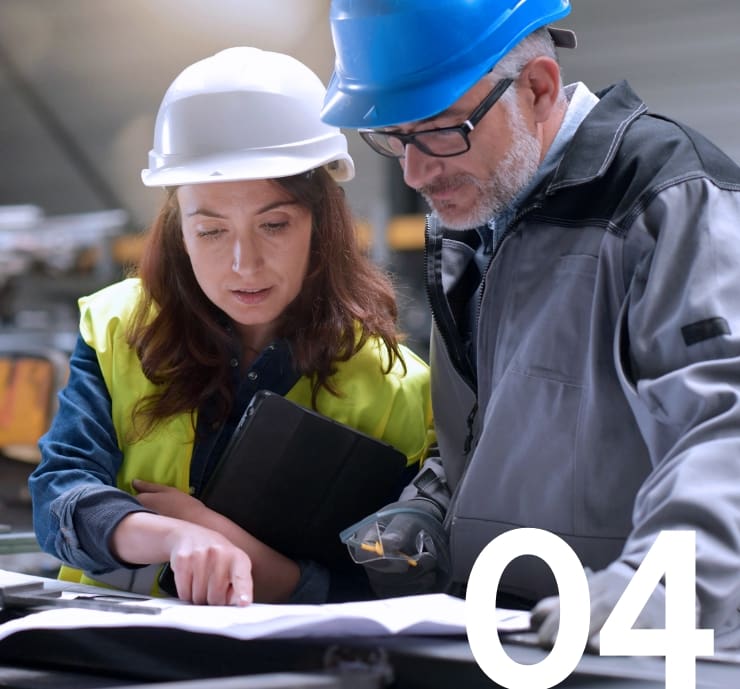 Two industrial engineers wearing hard hats and safety gear looking down reviewing technical plans.