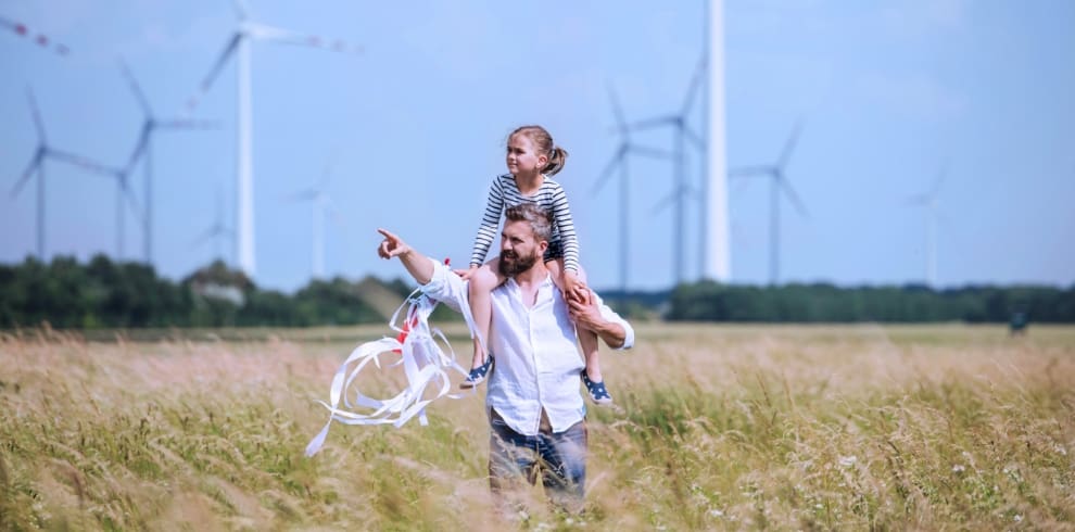 Father carrying child on shoulders in overgrown grassy field with win turbine farm in the background.