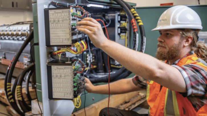 Electrical technician wearing a hard hat and safety vest working on industrial control panels with exposed wiring.