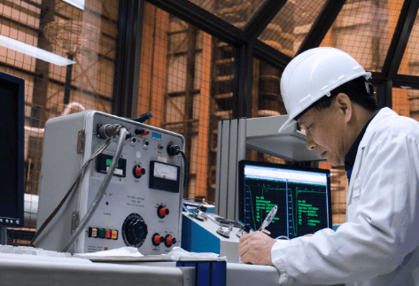 Engineer in a hard hat and lab coat conducting electrical equipment testing and analyzing data on condition monitoring, grid reliability, and failure analysis.